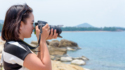 Young woman was happy to photography with dslr camera on the rock near the sea under the summer sky at Koh Samui island, Surat Thani province, Thailand