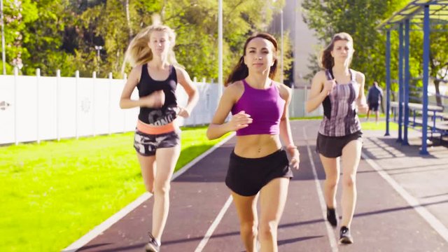 Dolly shot of three young women running along the path for runners at the sports ground. Front view