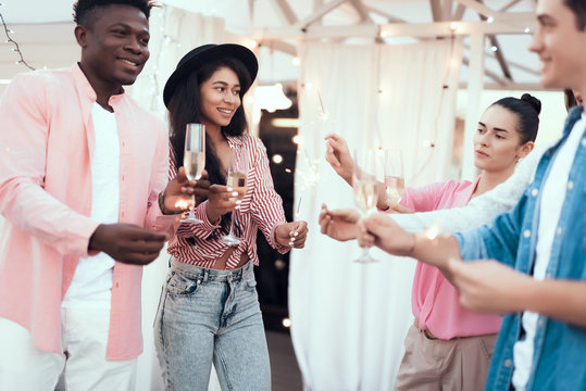 Outgoing girls holding firework sparklers while talking with cheerful men. They drinking glasses of champagne during party. They gesticulating hands