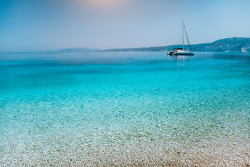 Obraz premium Sailing catamaran yacht boat at anchor near pebble beach with calm pure clear azure blue water surface