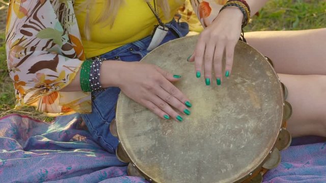 young blond hippie woman sitting on grass in park at sunset. Warm summer day and soft backlight. happy woman playing the tambourine and singing. slow motion HD