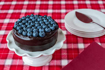 Round chocolate cake topped with fresh blueberries, on a white ceramic cake plate, red and white plaid tablecloth, red cloth napkin, silver cake server, stack of white dessert plates