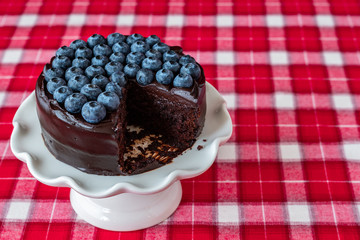Round chocolate cake topped with fresh blueberries, on a white ceramic cake plate, red and white plaid tablecloth, slice missing