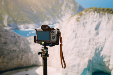 Digital photo camera on tripod against huge cliff rocks of Navagio beach in morning sun light. Famous visiting landmark location on Zakynthos island, Greece