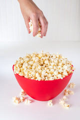 Vertical of hand reaching for popped popcorn in a big red plastic bowl on a white table, with spilled kernels