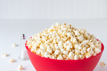 Popped popcorn in a big red plastic bowl on a white table, with a glass salt shaker and spilled kernels