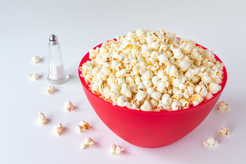 Popped popcorn in a big red plastic bowl on a white table, with a glass salt shaker and spilled kernels