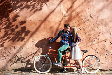 Young attractive happy romantic tourist couple in casual clothing, bearded man and blond woman in glasses standing together on double tandem bike on background of high red old crackled plastered wall.