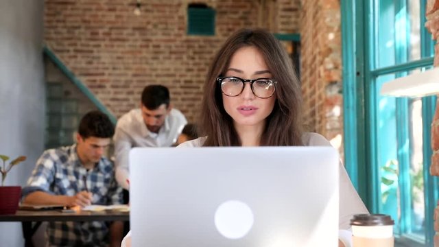 Young Attractive Woman Working on her Computer. Looking at Display. Wearing Eyeglasses.Surfing the Internet. Typing Mail Messages.Working on Start up. Group of Business People in the Background.