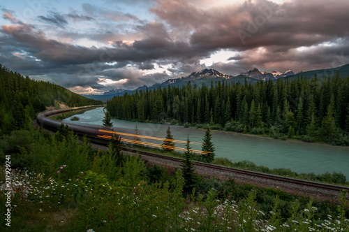 Train Rushing By River And Forest With Rocky Mountains In