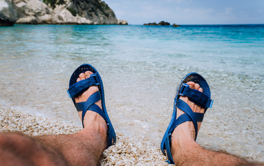 Young male feet wear blue flip-flop sandal sunbathing on pebble beach in front of blue sea water and rocks in background on horizon