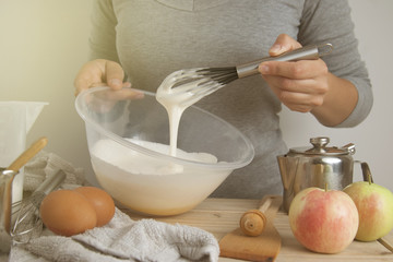 Close-up young woman's hands holding whisk and bowl while making pie, cake. Female cooking dough for pie on wooden table. Preparring dessert.