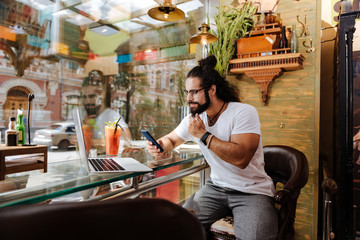 Coffee place. Confident brunette man using his gadgets while sitting in front of the window in the cafe