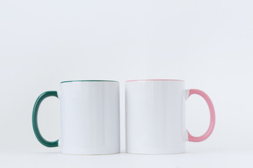 Two white mugs, with a green and pink handle on a light background. Side view.