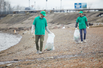 Activists in green uniform walking on shore and cleaning it from garbage