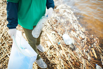 Crop unrecognizable activist in green uniform putting garbage to plastic bag on shore