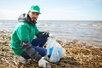 Young volunteer looking at camera and putting rubbish to plastic bag while sitting on shore - Powered by Adobe