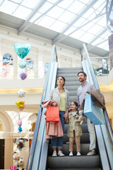 Parents with little girl carrying bags and moving on escalator in light big shopping center...