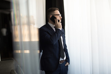 Serious stylish classy man in glasses having call on smartphone while standing and looking away in window