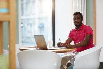 Adult casual African-American man sitting at table against window with laptop and cup of coffee taking notes and looking at camera