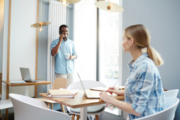 Obraz premium Side view of young lady with laptop sitting at office table and looking at African-American colleague talking on smartphone