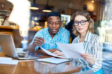 African-American man and woman in glasses sitting at table and reading documents together during business meeting in cafe