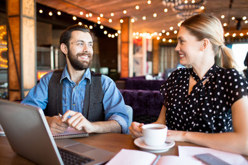 Trendy man and woman sitting at table with notes and laptop and working cheerfully having coffee in cafeteria