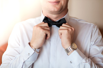 The groom man in a white shirt straightens his hands with a black bow tie. Close up