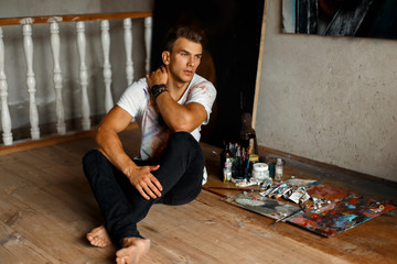 Stylish handsome young man artist in a white tank top with paint sits on the floor in the studio....