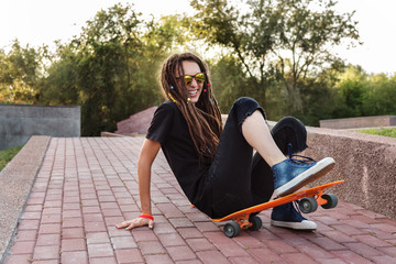fashionable girl with dreadlocks in sunglasses fell from the skateboard and laughs. positive girl...