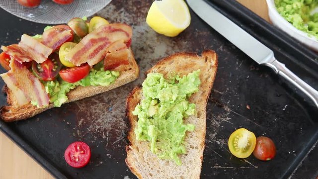 A woman puts mashed avocado, cherry tomatoes and bacon on toasted bread
