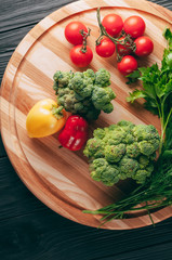 Fresh vegetables: cherry tomatoes, broccoli, pepper, dill, parsley on a round wooden board, on a dark wooden table. Recipe. Ingredients. Dietary food. Place under the text. View from above.