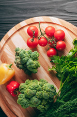 Fresh vegetables: cherry tomatoes, broccoli, pepper, dill, parsley on a round wooden board, on a dark wooden table. Recipe. Ingredients. Dietary food. Place under the text. View from above.