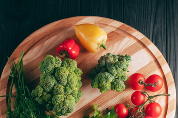 Fresh vegetables: cherry tomatoes, broccoli, pepper, dill, parsley on a round wooden board, on a dark wooden table. Recipe. Ingredients. Dietary food. Place under the text. View from above.