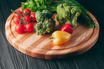 Fresh vegetables: cherry tomatoes, broccoli, pepper, dill, parsley on a round wooden board, on a dark wooden table. Recipe. Ingredients. Dietary food. Place under the text. View from above.