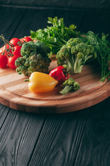 Fresh vegetables: cherry tomatoes, broccoli, pepper, dill, parsley on a round wooden board, on a dark wooden table. Recipe. Ingredients. Dietary food. Place under the text. View from above.
