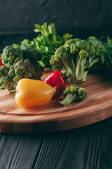 Fresh vegetables: cherry tomatoes, broccoli, pepper, dill, parsley on a round wooden board, on a dark wooden table. Recipe. Ingredients. Dietary food. Place under the text. View from above.
