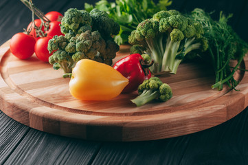 Fresh vegetables: cherry tomatoes, broccoli, pepper, dill, parsley on a round wooden board, on a dark wooden table. Recipe. Ingredients. Dietary food. Place under the text. View from above.