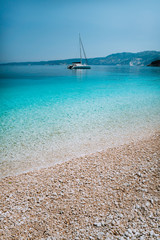 Pebble beach with pure clear azure blue water and sailing catamaran yacht boat at anchor on horizon