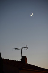 Fototapeta na wymiar La lune et une étoile brillent au-dessus d'un toit d'où se dresse une antenne de télévision.