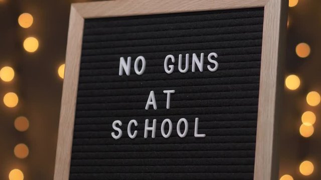 Black letter board with NO GUNS AT SCHOOL Written on it with white letters. Camera rotating around the sign showing the beautiful bokeh balls in the background.