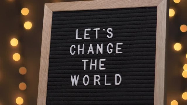 Black letter board with LET'S CHANGE THE WORLD Written on it with white letters. Camera rotating around the sign showing the beautiful bokeh balls in the background.