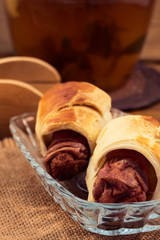 Fried meat sausages baked in puff pastry until golden  brown in a glass plate on the background of a jug of herbal tea, linen napkin, wooden kitchen tongs.