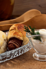 Puff roll of dough with meat sausages, baked in the oven until brown, sour cream in a gravy boat with a sprig of dill, linen napkin, wooden kitchen tongs, on the background of a jug of tea from herbs.