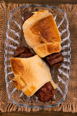 Fried meat sausages baked in puff pastry until golden  brown in a glass plate on a linen cloth napkin, top view on a wooden background.
