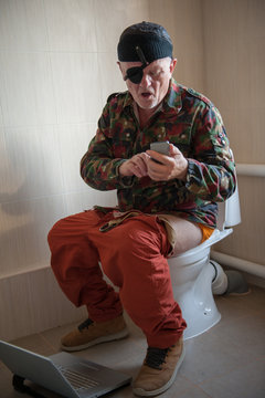 Man with gray hair, beard and mustache in military jacket with laptop sitting on toilet. Man's eye is covered with black medical patch. Dude is infatuated with computer and actively expressed emotions