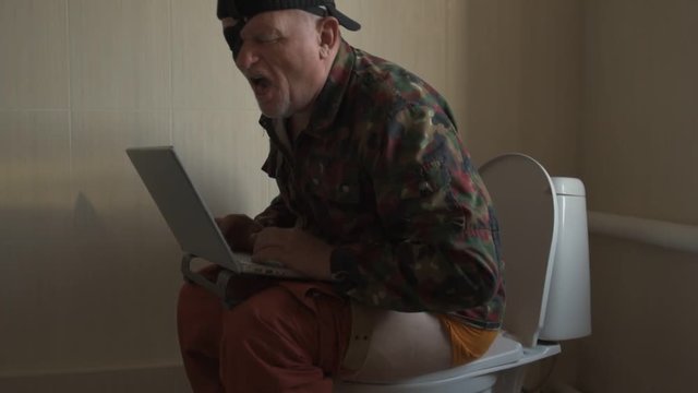 Man with gray hair, beard and mustache in military jacket with laptop sitting on toilet. Man's eye is covered with black medical patch. Dude is infatuated with computer and actively expressed emotions