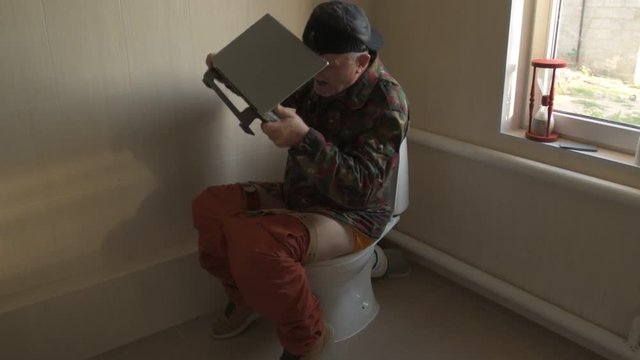 Man with gray hair, beard and mustache in military jacket with laptop sitting on toilet. Man's eye is covered with black medical patch. Dude is infatuated with computer and actively expressed emotions