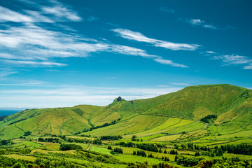 A mountain with green fields and a blue sky with some clouds