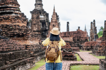 Fototapeta premium Asian tourist woman take a photo of ancient of pagoda temple thai architecture at Sukhothai Historical Park,Thailand. Female traveler in casual thai cloths style visiting city concept.